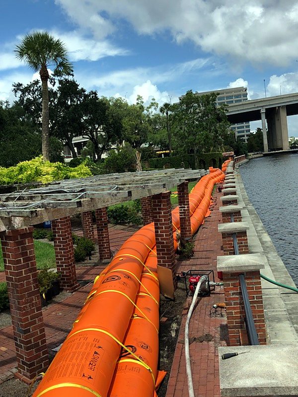 Stacked water-filled barrier tubes along a brick museum promenade beside a river