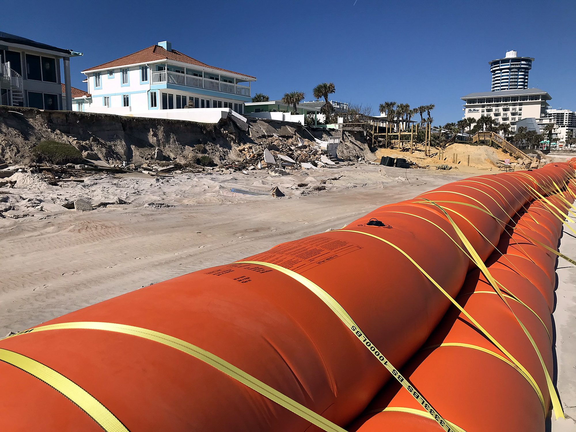 Long stacked orange water-filled barrier running along an oceanfront beach below eroded dunes