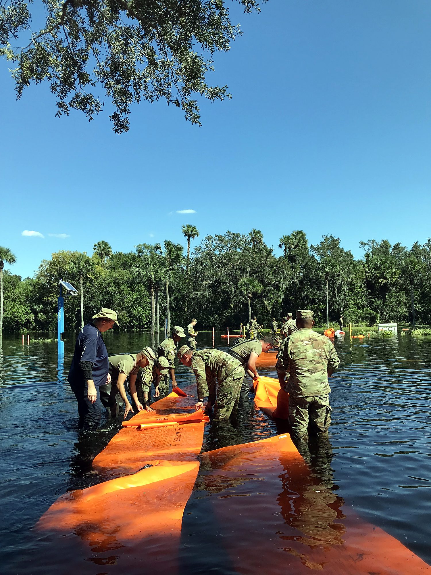 National Guard personnel in uniform working a water-filled barrier line in knee-deep floodwater