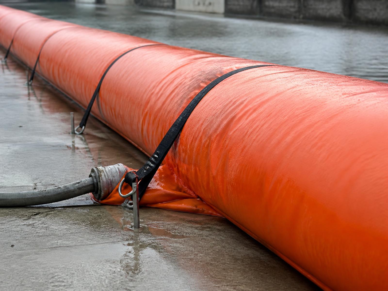 Close view of a water-filled barrier tube with anchoring straps, stakes and a fill hose on wet concrete